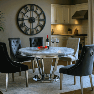 Amodini round dining table with grey marble top and polished steel pedestal base, surrounded by tufted dining chairs in a classic dining room.